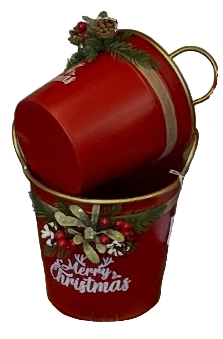 A red Christmas-themed metal bucket with gold trim and a hanging white tag, filled with a matching red bucket tipped on its side. The bucket features green pine garland, red berries, and white 'Merry Christmas' lettering.