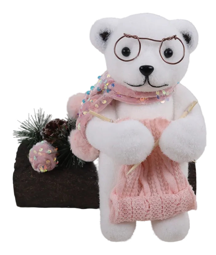 A pink bear wearing glasses, a sparkling pink scarf, and knitting a hat, seated next to a decorative log.