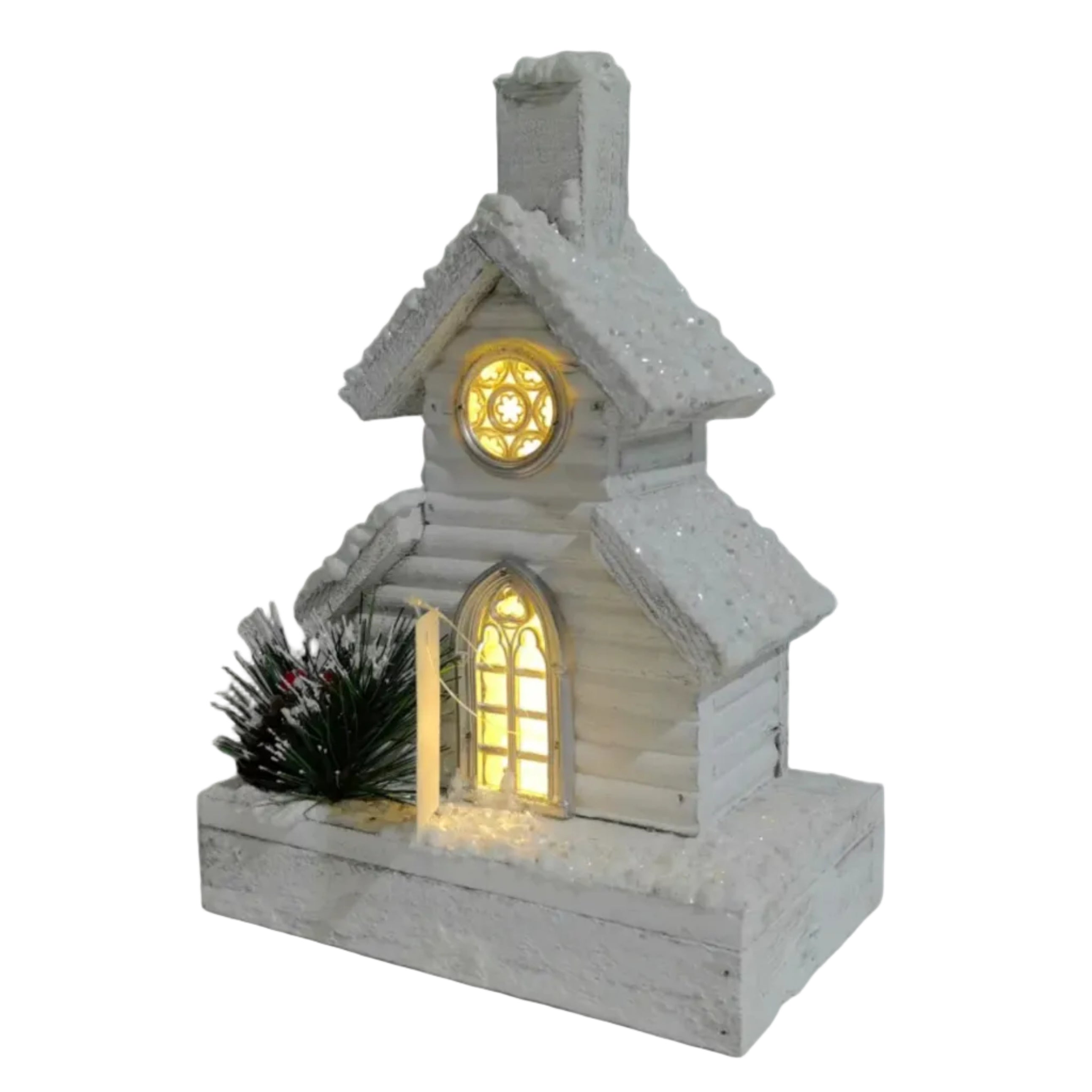 A small white snow-covered chapel Christmas ornament with warm yellow glowing lights in the windows and a tiny green pine tree on the porch.