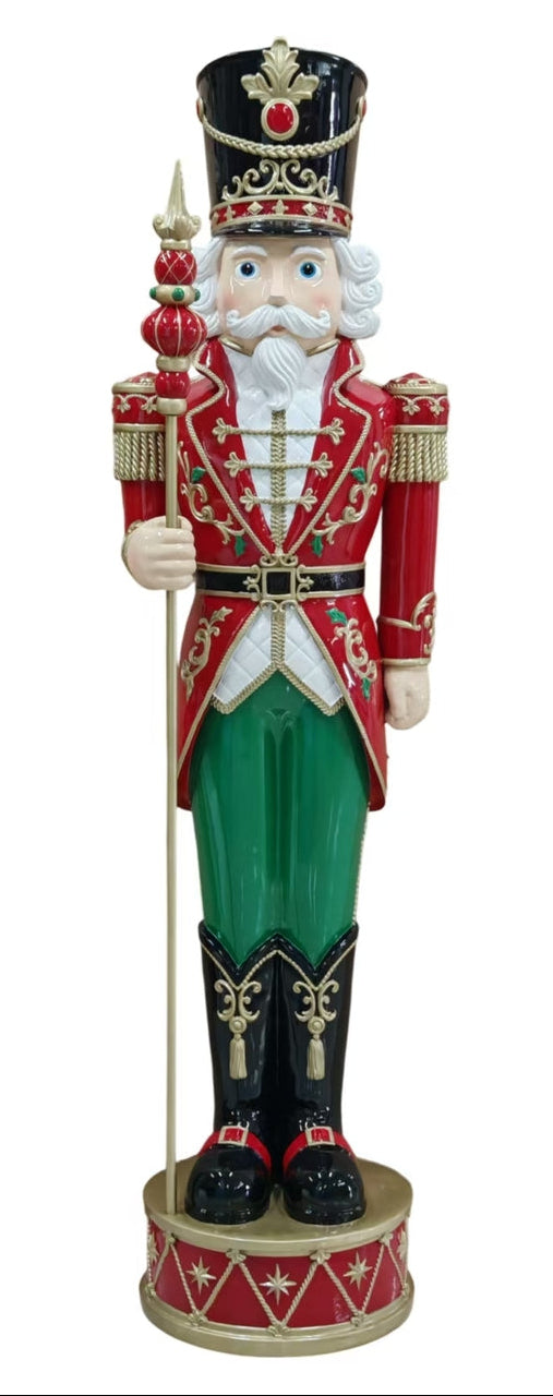 A traditional Christmas nutcracker figurine standing on a drum-shaped base, wearing a red coat, green pants, black boots, and a black hat with gold details, holding a spear.