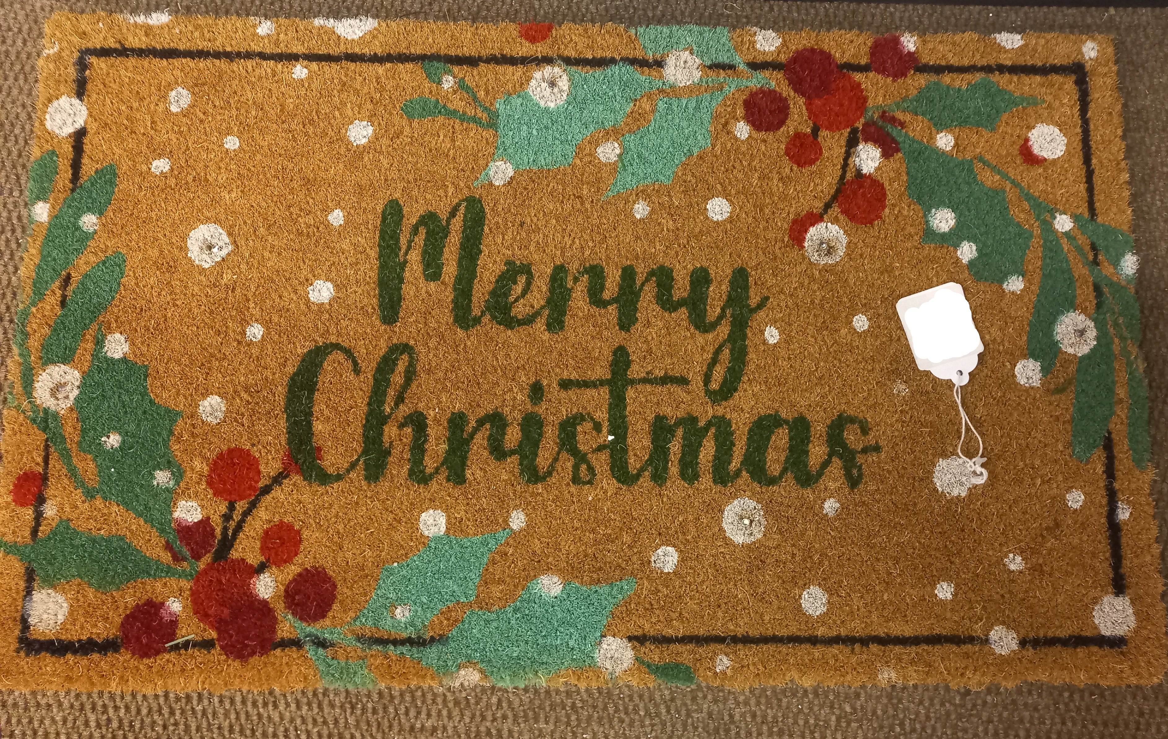 A rectangular coir doormat with a brown natural fiber base, featuring a green 'Merry Christmas' script, red and green holly leaves, white snowflake motifs, and a black border. 