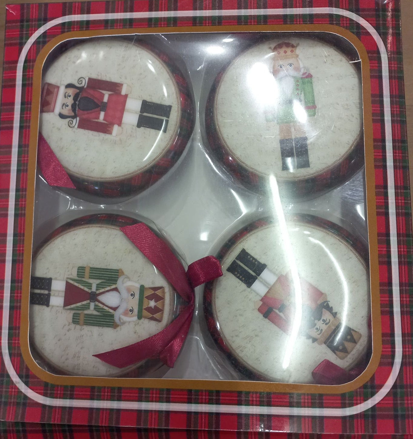 Packaged set of four round flat nutcrackers in a clear plastic box with a red and black plaid design and decorative ribbons.